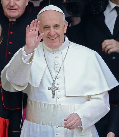 Un hombre en vestimenta religiosa blanca con una cruz prominente en el pecho, saluda sonriendo desde un balc&oacute;n.