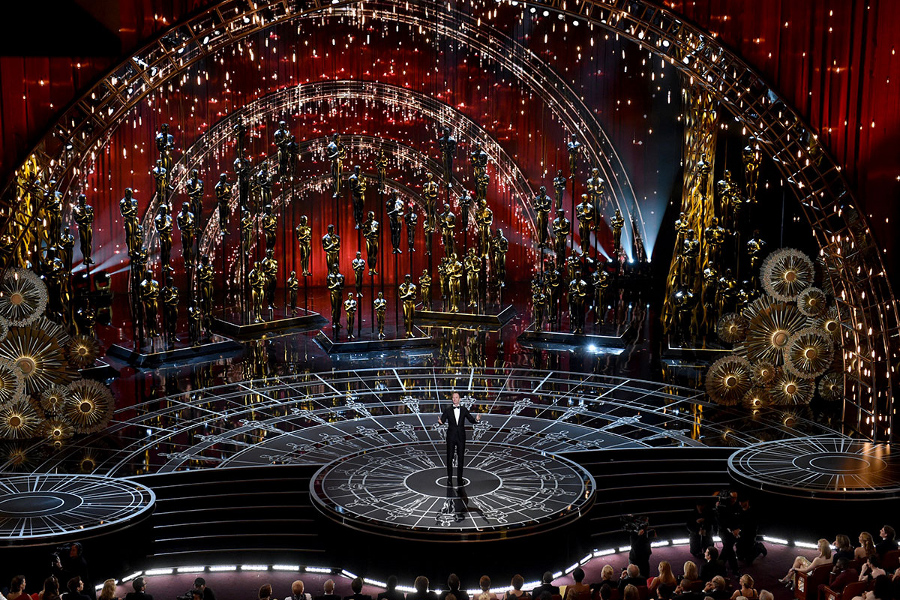 Una imagen de un gran escenario circular brillando con m&uacute;ltiples luces y decorado con esferas gigantes y figuras humanas doradas, evocando constelaciones y los Mazzaroth. Un hombre vestido de traje se encuentra en el centro, hablando frente a una audiencia en un teatro repleto.
