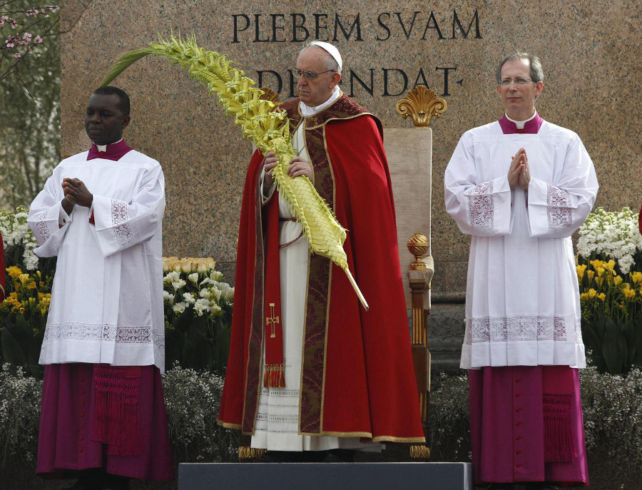 Tres cl&eacute;rigos durante una ceremonia religiosa al aire libre. A la derecha, un cl&eacute;rigo sostiene una larga palma, vestido con ornamentos rojos y blancos. A la izquierda, dos cl&eacute;rigos en vestiduras blancas y p&uacute;rpuras est&aacute;n de pie, uno con las manos juntas en posici&oacute;n de oraci&oacute;n. Al fondo, flores y un monumento con una inscripci&oacute;n en lat&iacute;n.