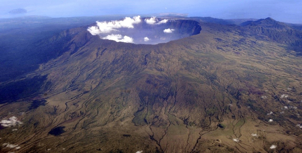 Vista a&eacute;rea de un extenso cr&aacute;ter volc&aacute;nico rodeado de terreno monta&ntilde;oso, con nubes form&aacute;ndose sobre el cr&aacute;ter, resaltando la majestuosidad de la creaci&oacute;n que se extiende bajo el firmamento.