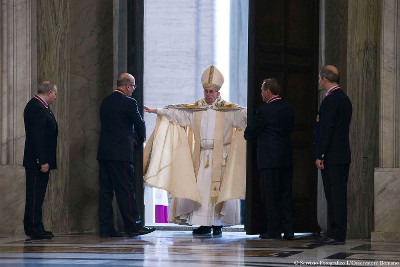 Un l&iacute;der religioso vestido con una t&uacute;nica ceremonial alta y mitra, extendiendo sus brazos en un gesto amplio, rodeado de varios hombres de traje en un entorno de columnas de m&aacute;rmol.